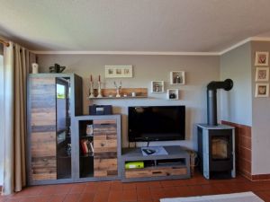 Wohnzimmer Offene Küche. Wohnzimmer mit Schlafcouch für zwei Personen und Couchtisch, Wohnzimmerwand, Esstisch mit Sitzbank und drei Stühlen, Kaminofen. Zur Unterhaltung stehen ein Flachbild-TV mit Sat-Empfang, ein DVD-Player und ein Radio zur Verfügung.