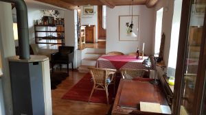 Wohnzimmer Richtung Küche Rechte Fensterseite: Antik-Schreibtisch mit Hocker, Esstisch. Linke Seite: Kaminofen (daneben noch Katalytofen), Schlafsofa (verdeckt), Lederstuhl. Mitte: drei Stufen zum Küchenbereich, Tür dahinter zur Abstellkammer